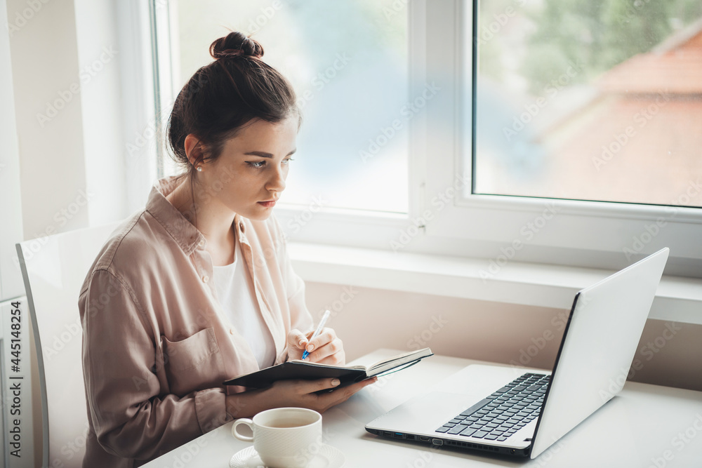 Brunette woman is writing in a copybook something while looking at the laptop screen during workday at the office Brunette woman is writing in a copybook something while looking at the laptop screen during workday at the office