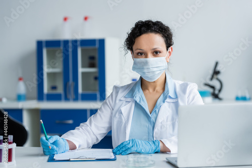 African american doctor in medical mask and latex gloves sitting near clipboard and laptop in lab