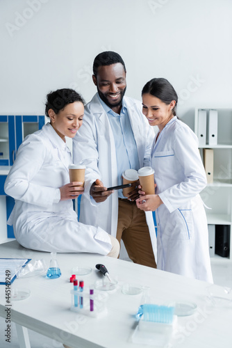 Positive interracial scientists with coffee looking at smartphone in laboratory