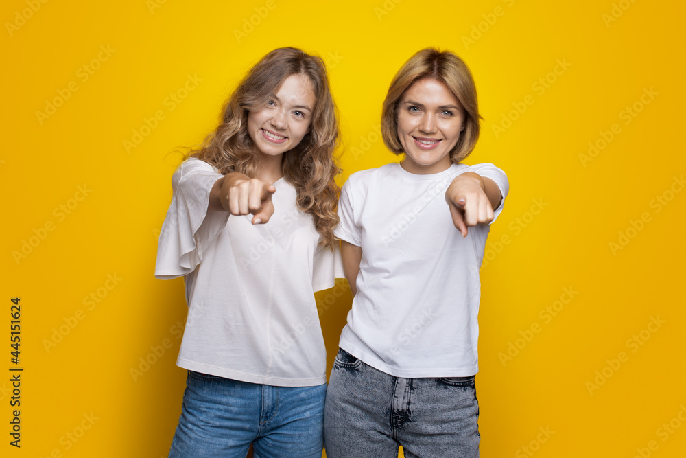 Blonde haired sisters are pointing at camera smiling on a yellow studio wall wearing t-shirt and jeans Blonde haired sisters are pointing at camera smiling on a yellow studio wall wearing t-shirt and jeans