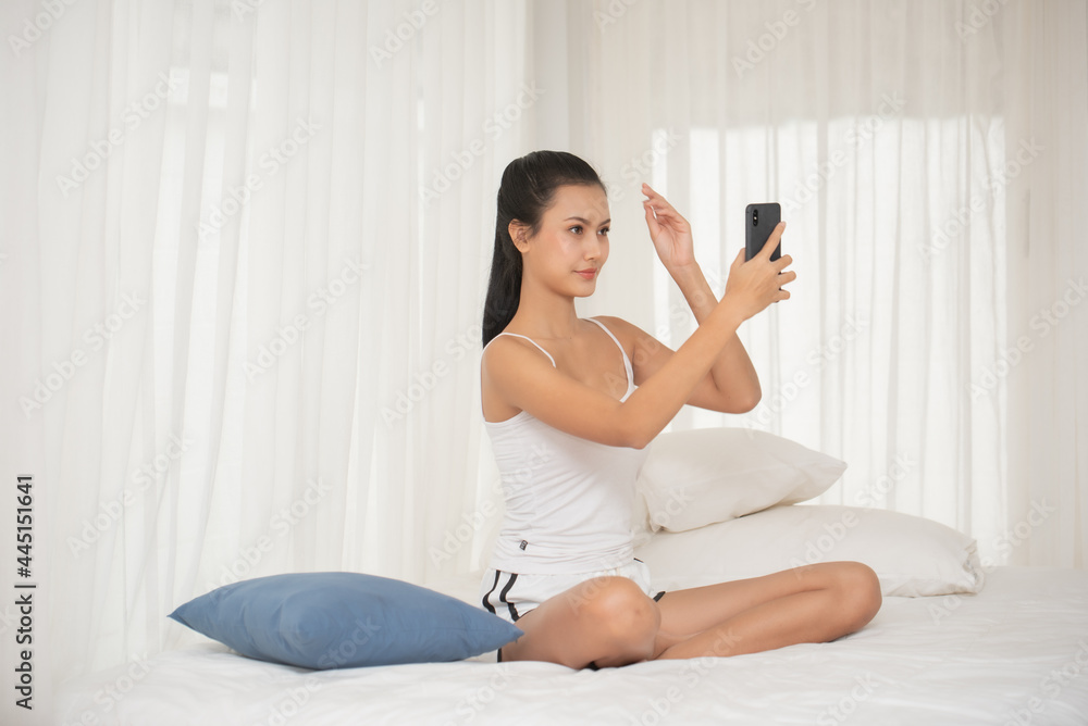 Young Woman Using Mobile Phone While Sitting On Bed At Home Young Woman Using Mobile Phone While Sitting On Bed At Home