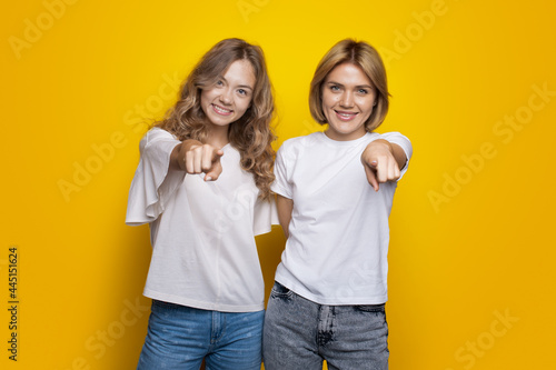 Blonde haired sisters are pointing at camera smiling on a yellow studio wall wearing t-shirt and jeans