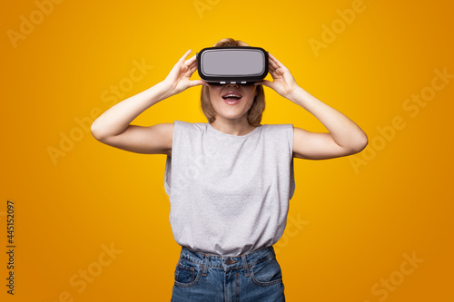 Adorable woman is smiling while using a virtual reality headset getting surprised on a yellow studio wall