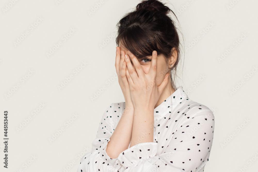 Adorable woman with brunette hair hiding herself with palms at camera wearing a white shirt on a studio wall Adorable woman with brunette hair hiding herself with palms at camera wearing a white shirt on a studio wall
