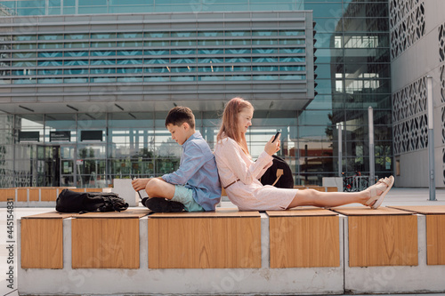 Portrait of cute young students wearing casual, sitting back to back, holding their mobile devise in hands, seriously looking at screens, schoolyard near campus