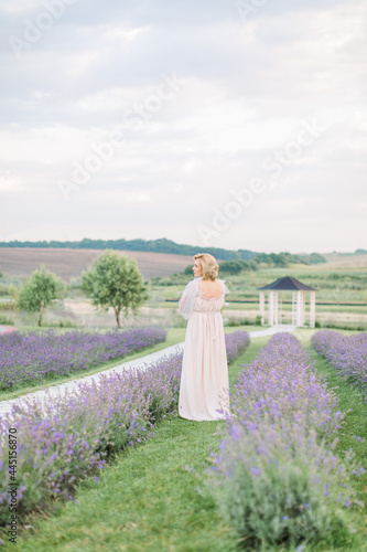 Beautiful middle aged gorgeous blond lady in elegant dress walking in the field of lavender. Back full length view.