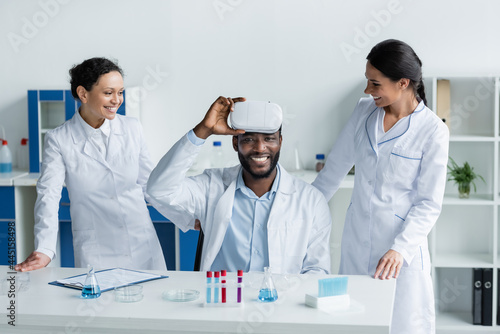 Cheerful african american scientist holding vr headset near colleagues in laboratory