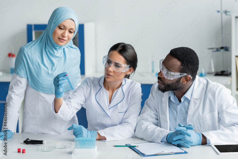 Scientist holding test tube near interracial colleagues in safety goggles, electronic pipette and digital tablet Scientist holding test tube near interracial colleagues in safety goggles, electronic pipette and digital tablet