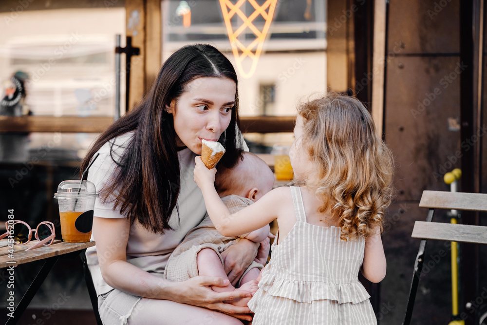 Caucasian toddler girl feeding baby brother boy with ice cream cone. Mother is laughing. Summer family fun outdoors. Sister teasing child with forbidden food Caucasian toddler girl feeding baby brother boy with ice cream cone. Mother is laughing. Summer family fun outdoors. Sister teasing child with forbidden food