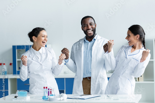 Excited interracial scientists showing yes gesture near smartphone, clipboard and test tubes