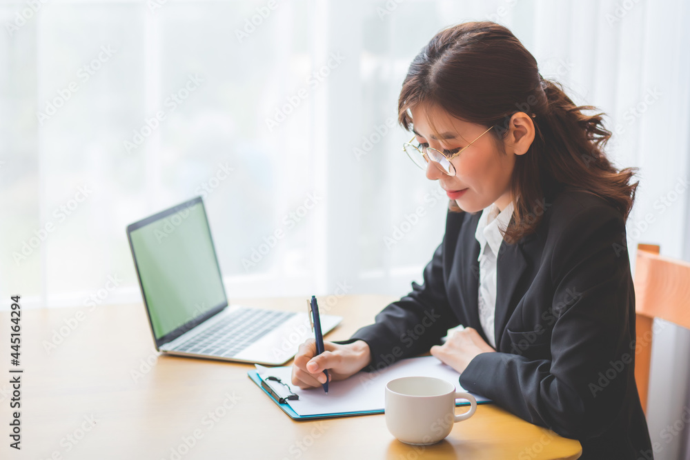 A beautiful Asian woman in formal attire holding a pen and writing or taking notes on a table with a laptop and coffee mugs placed in the concept of a work and online meeting from home. A beautiful Asian woman in formal attire holding a pen and writing or taking notes on a table with a laptop and coffee mugs placed in the concept of a work and online meeting from home.