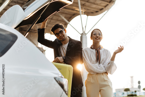 Brunette man in black suit and blonde attractive woman in white outfit look into camera. Guy puts yellow luggage into car trunk. Lady in eyeglasses smiles widely outside. © Look!