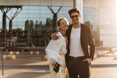 Brunette happy man in white shirt and black suit hugs attractive girlfriend near airport. Charming lady in white blouse and eyeglasses smiles and holds passport.