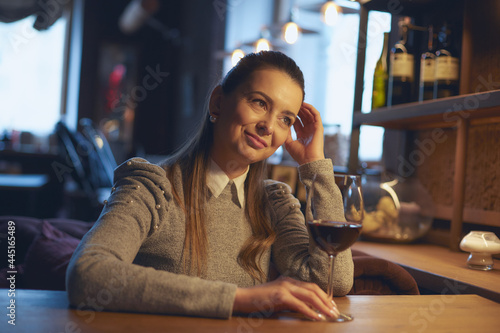 A beautiful young woman sits at a wooden table in the premises of a wine bar and drinks wine from a glass.