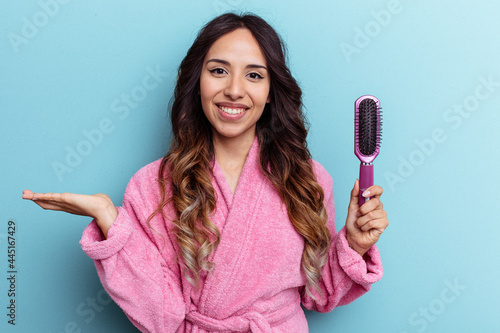 Young mexican woman wearing a bathrobe holding a brush isolated on blue background showing a copy space on a palm and holding another hand on waist.