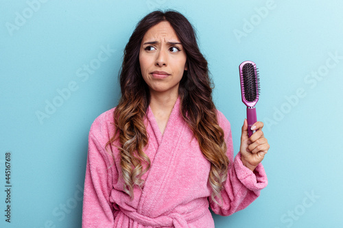 Young mexican woman wearing a bathrobe holding a brush isolated on blue background confused, feels doubtful and unsure.