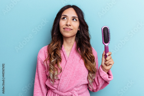 Young mexican woman wearing a bathrobe holding a brush isolated on blue background dreaming of achieving goals and purposes