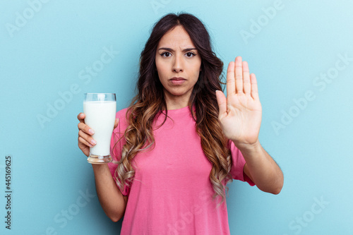 Young mixed race woman holding a glass of milk isolated on blue background standing with outstretched hand showing stop sign, preventing you.