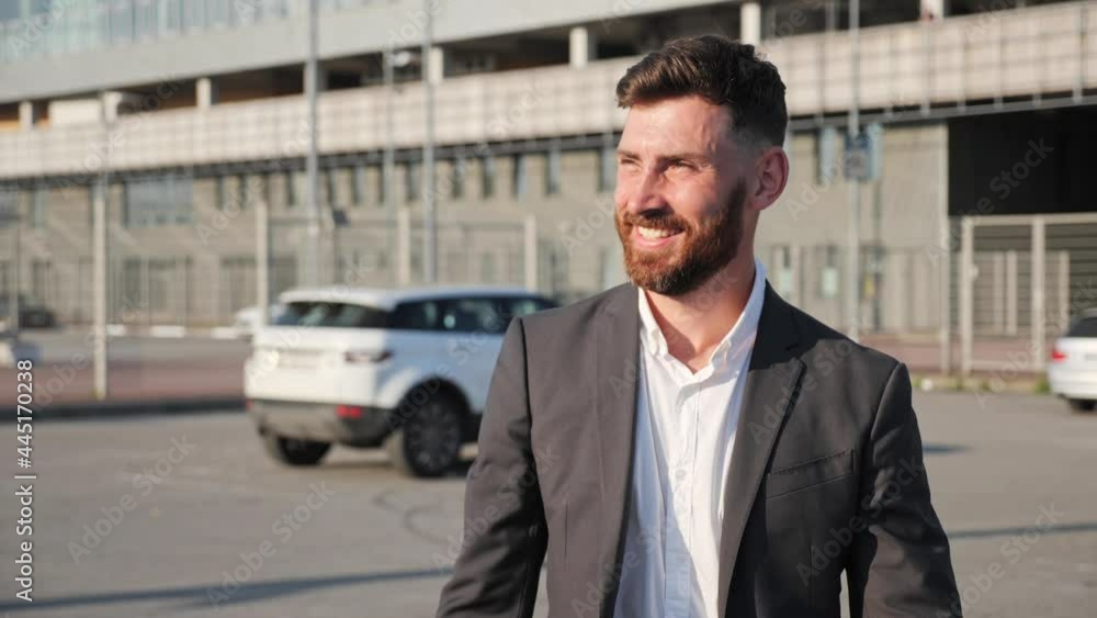 Smiling bearded businessman dressed in classic suit is walking through the parking. Young millennial man waving to friend near white car on the background. Business. Successful people concept. Smiling bearded businessman dressed in classic suit is walking through the parking. Young millennial man waving to friend near white car on the background. Business. Successful people concept.