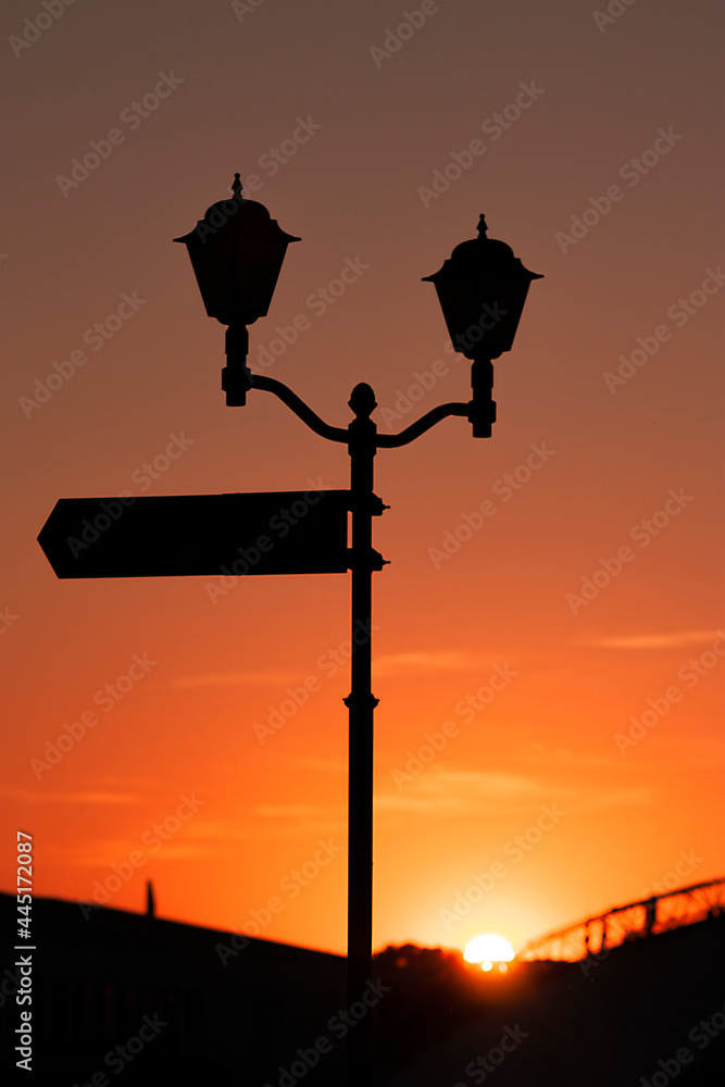 Vertical photography with a silhouette of a vintage street lamps and way sign on the pole, against orange sky at sunset