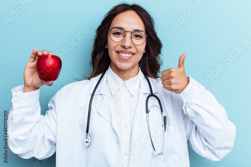 Young doctor latin woman holding an apple isolated on blue background
