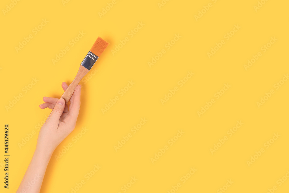 Female hand hold paintbrush on a yellow background. Repair concept with copyspace. Female hand hold paintbrush on a yellow background. Repair concept with copyspace.