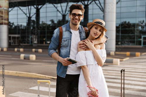 Attractive blonde woman in hat, white dress holds pink camera. Handsome brunette man in denim jacket hugs girlfriend and holds passport near airport. © Look!