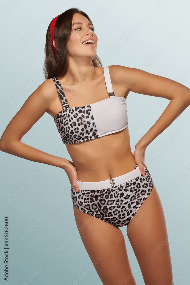 Fototapeta premium Slim lady is wearing two-piece leopard swimsuit composed of bra with shoulder straps and high-waisted panties. Smiling girl with red headband is posing on the blue background.
