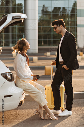 Woman in white blouse, beige pants sits in car trunk and talks to her boyfriend. Brunette man and blonde lady speak and pose with yellow suitcase near airport.