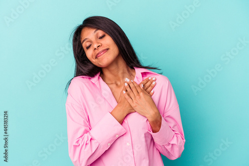Young latin woman isolated on blue background has friendly expression, pressing palm to chest. Love concept.