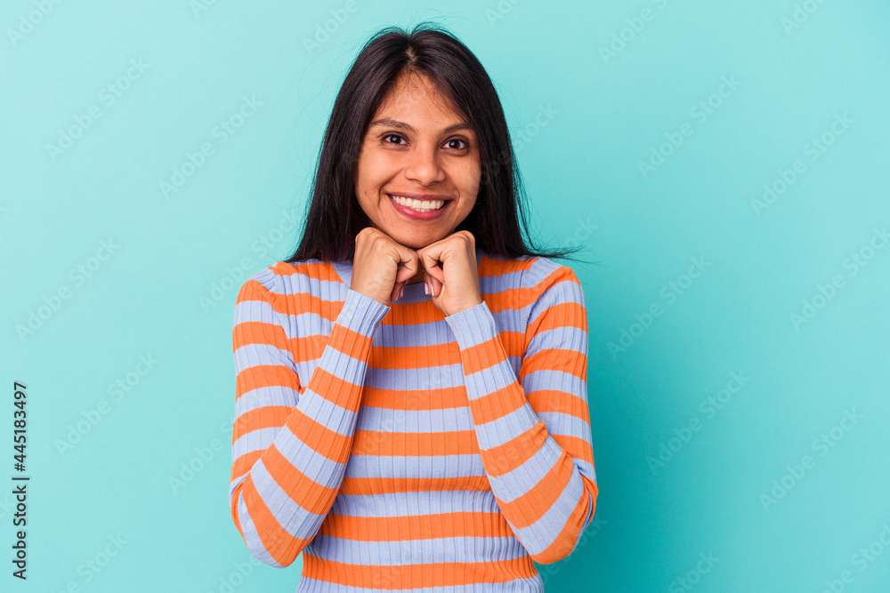 Young latin woman isolated on blue background keeps hands under chin, is looking happily aside. Young latin woman isolated on blue background keeps hands under chin, is looking happily aside.