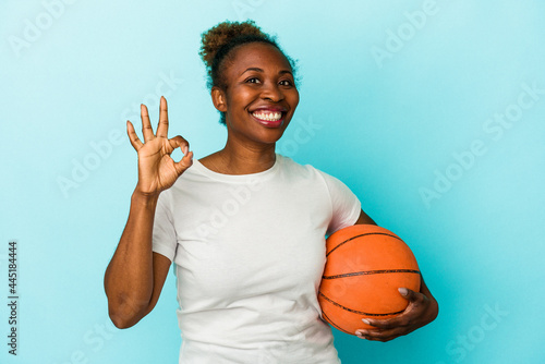 Young african american woman playing basketball isolated on blue background cheerful and confident showing ok gesture. © Asier