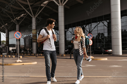 Full-length portrait of curly blonde woman and man in jeans and white tees walking near airport. Travelers talking and posing with backpacks.