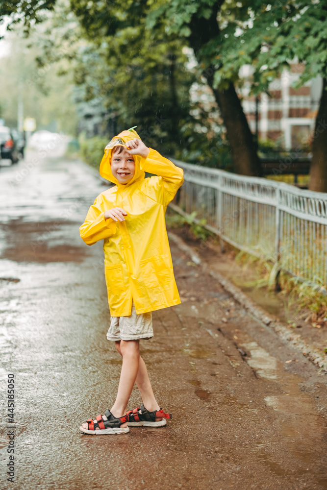 cute boy in a yellow raincoat and sandals is having fun, running through the puddles, in the city. jump through muddy puddles. Happy and carefree childhood