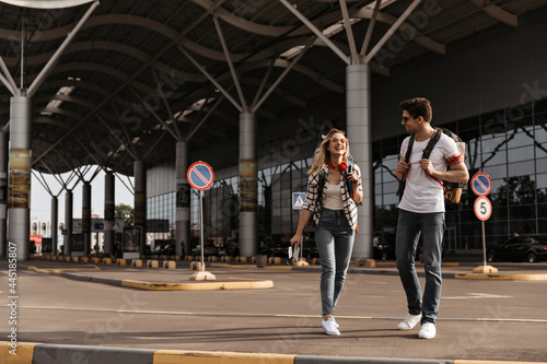 Happy blonde woman talks with her boyfriend. Full-length portrait of cool girl in red headphones and brunette guy in white tee walks with backpacks near airport.