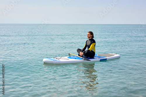 Side view portrait of relaxed bearded guy sitting on paddleboard meditating after sup surfing, in black wetsuit, keep calm, yoga and sport concept. copy space
