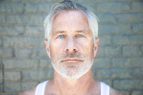 Fit Man with grey hair and beard blue eyes wearing white tank top facing camera with grey brick backdrop