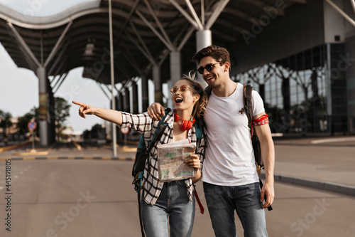 Happy girl points at left side and smiles. Man in white t-shirt hugs his blonde girlfriend. Couple of travelers hold map and backpacks near airport. © Look!