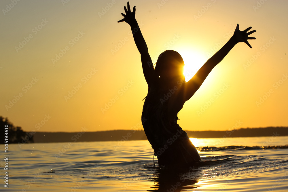 Silhouette of happy teenage girl standing a confident pose in a river, pond, lake water at sunset with hands in the air, fingers keeping apart. Summer vacations, holidays on a coast. Happy childhood.