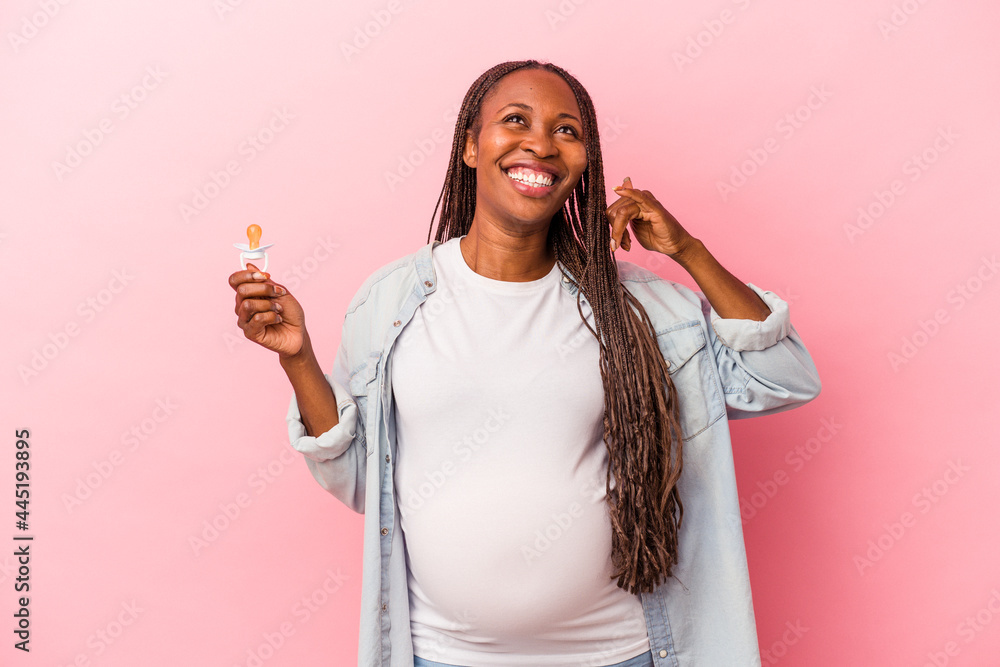 Young african american pregnant woman holding pacifier isolated on pink background showing a mobile phone call gesture with fingers. Young african american pregnant woman holding pacifier isolated on pink background showing a mobile phone call gesture with fingers.