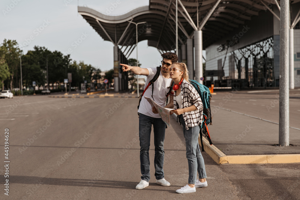 Tourists in jeans poses with map and decide where to go. Blonde woman and brunette man in white t-shirts and sunglasses talks near airport and holds backpacks.