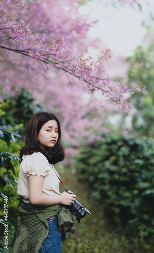 Asian teenage girl with a camera stands looking at it under a cherry blossom tree.