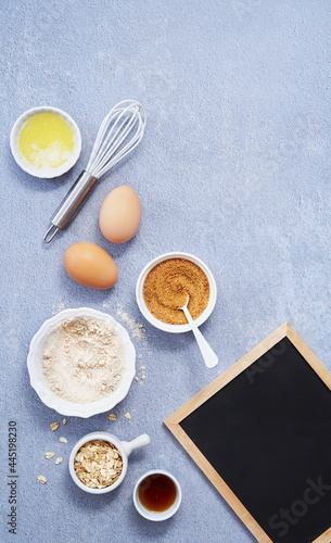 Food Ingredients, whole grain oat, coconut sugar, vanilla syrup, organic eggs next to black chalkboard on light blue surface. Healthy food recipe. Top view. Copy Space.