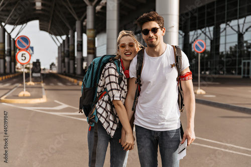 Couple of travelers poses near airport and looks into camera. Blonde woman in plaid shirt  sunglasses and brunette handsome man in white tee hold hands and poses with backpacks.