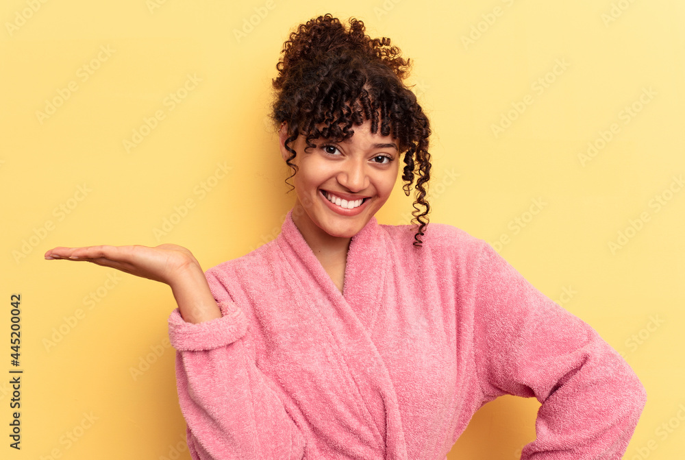Young mixed race woman wearing a pink bathrobe isolated on pink background showing a copy space on a palm and holding another hand on waist. Young mixed race woman wearing a pink bathrobe isolated on pink background showing a copy space on a palm and holding another hand on waist.