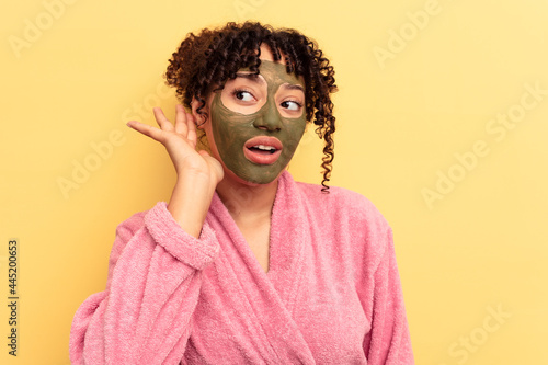 Young mixed race wearing facial mask isolated on yellow background trying to listening a gossip.