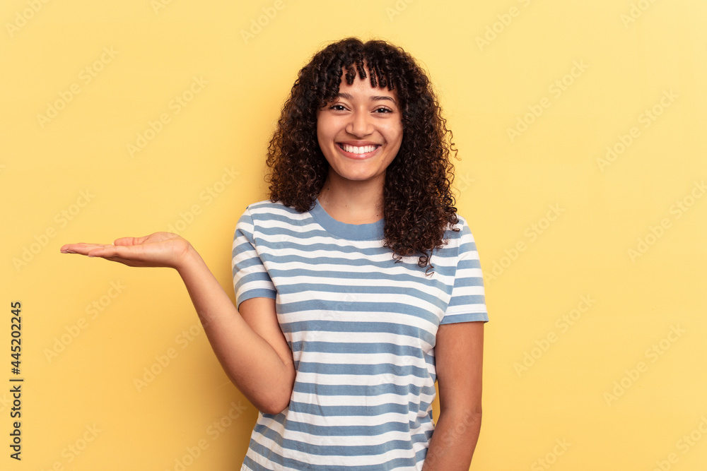 Young mixed race woman isolated on yellow background showing a copy space on a palm and holding another hand on waist. Young mixed race woman isolated on yellow background showing a copy space on a palm and holding another hand on waist.
