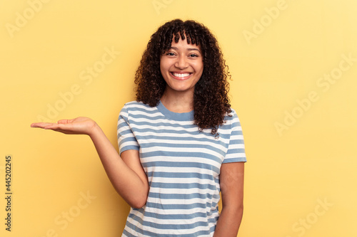 Young mixed race woman isolated on yellow background showing a copy space on a palm and holding another hand on waist. © Asier