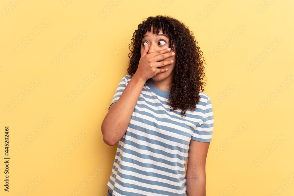 Young mixed race woman isolated on yellow background thoughtful looking to a copy space covering mouth with hand. Young mixed race woman isolated on yellow background thoughtful looking to a copy space covering mouth with hand.