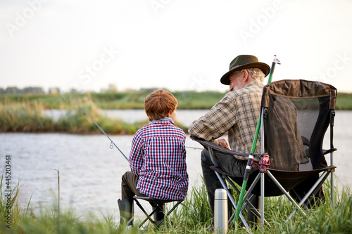 Canvas Print Rear view on senior man with grandson sitting near river with fishing rods in hands, enjoying beautiful nature, little boy learn to fish with grandfather